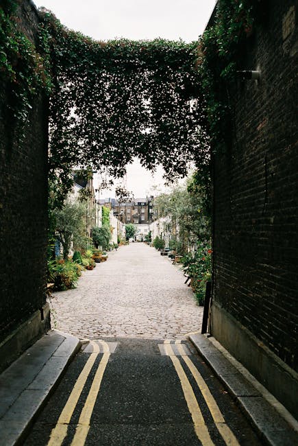 A narrow cobblestone alleyway viewed from a tunnel-like entrance framed by dark brick walls covered in lush, green ivy. The alley is lined with small potted plants and shrubs on both sides, adding vib