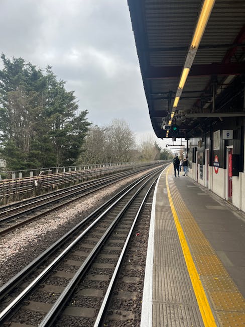 This image shows a section of a train platform at Rayners Lane station, with railway tracks running parallel to the platform. The platform surface is made of concrete, featuring a yellow tactile pavin