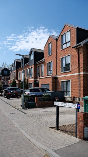 A row of modern, multi-story residential buildings constructed with red brick and black cladding, featuring large windows and pitched roofs under a bright blue sky with scattered clouds. In front of t